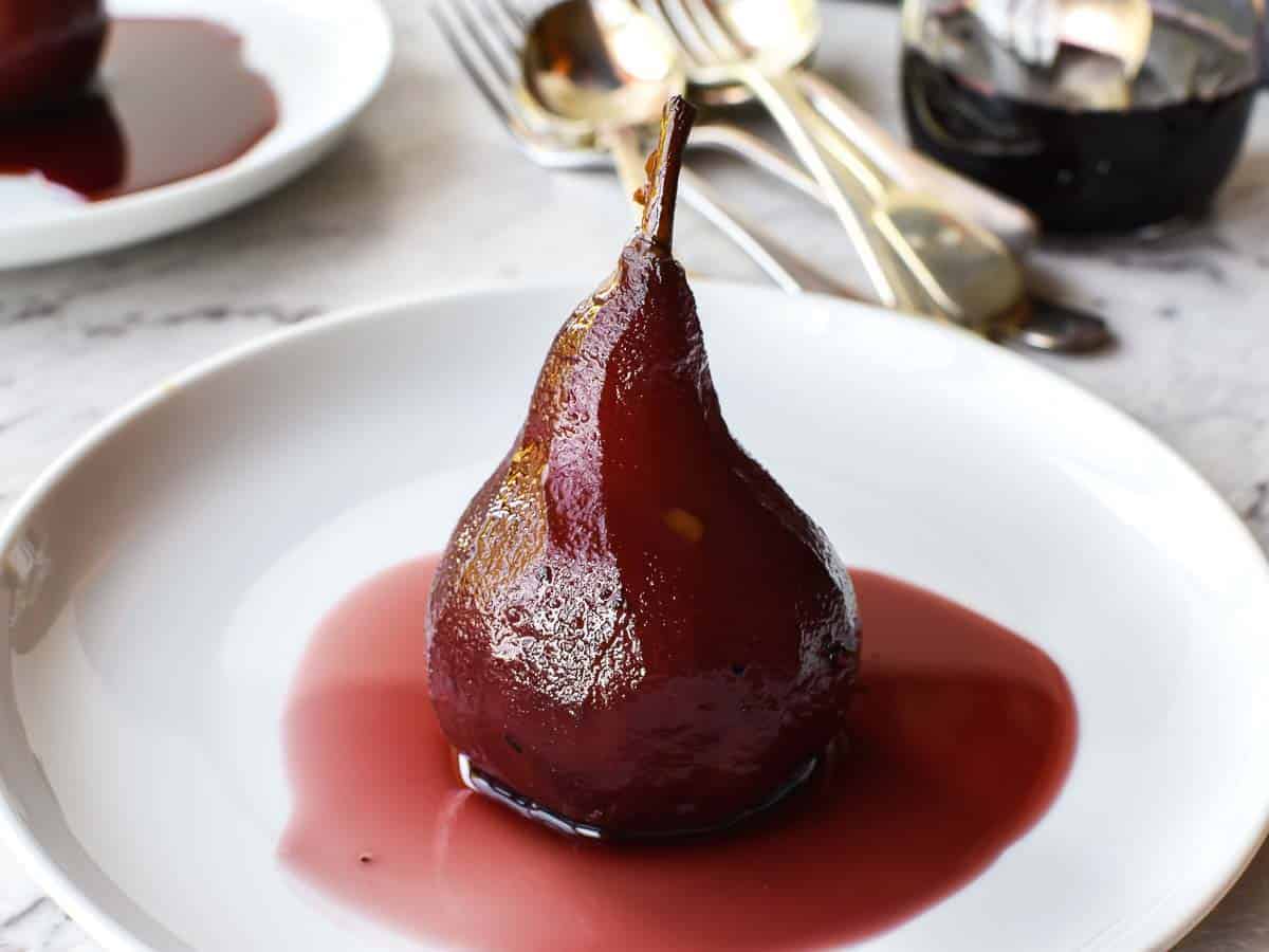 Poached Pears in Red Wine and Spices is a simple and impressive dessert that is equally at home at a dinner party or family meal. These delicious pears glisten with syrup and are fragrant with spices. Included is a special ingredient that balances out the sweetness. #pears #pearrecipe #redwine #dessert #recipe #poachedpears A poached pear sitting upright in red syrup on a white plate