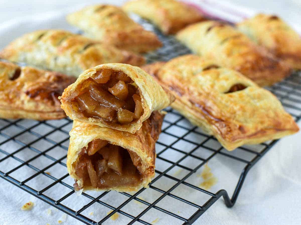 These homemade Apple Turnovers made with puff pastry are so easy. This recipe is really simple to make using store bought dough and a fresh apple pie filling. Enjoy bakery style, flaky pastries that you have baked yourself! #apple #appleturnovers #puffpastry #homemade #pies #pastry #applepies Pastries with apple filling on a black wire cooling rack with one cut in half to reveal the inside.