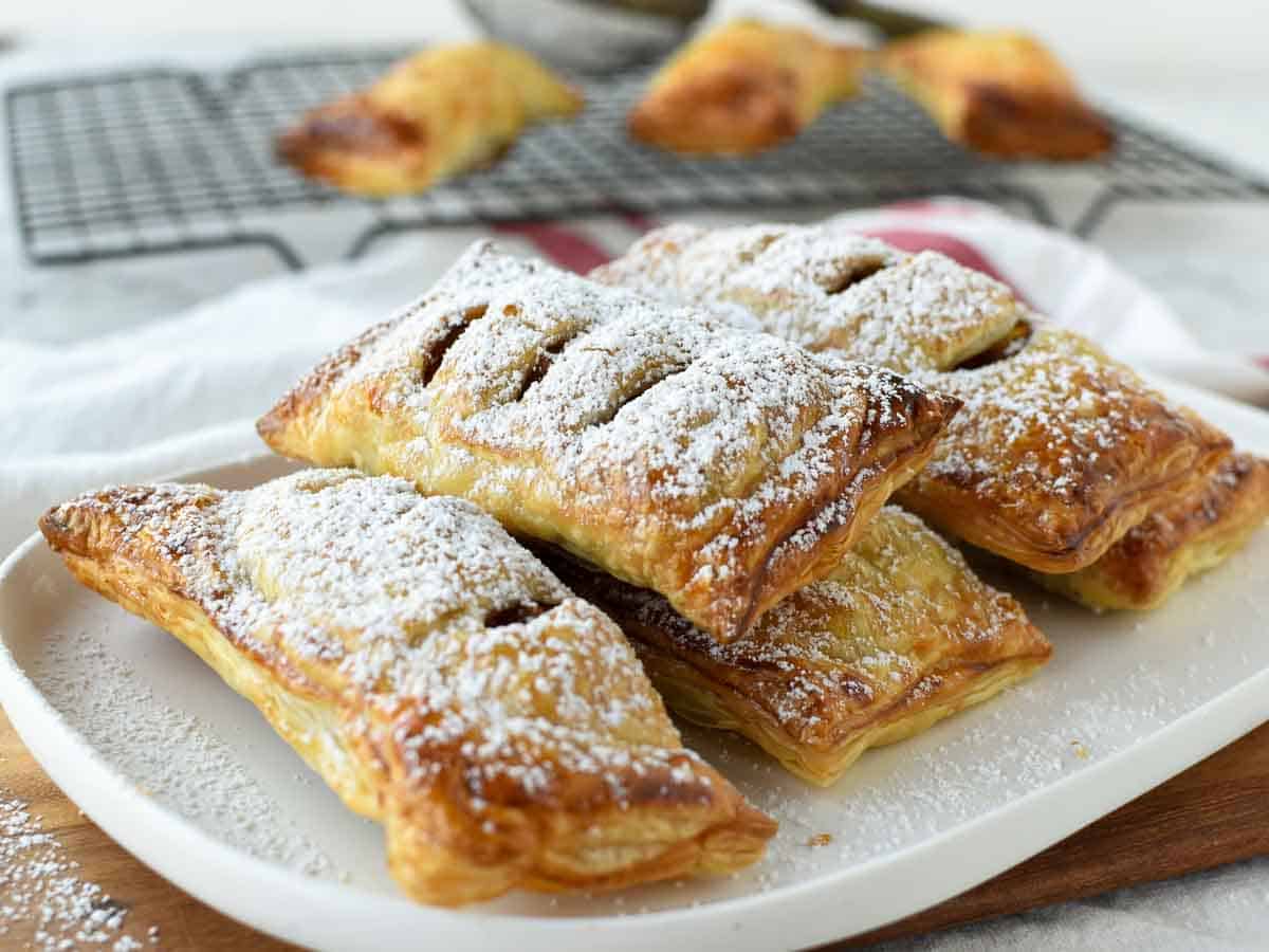 These homemade Apple Turnovers made with puff pastry are so easy. This recipe is really simple to make using store bought dough and a fresh apple pie filling. Enjoy bakery style, flaky pastries that you have baked yourself! #apple #appleturnovers #puffpastry #homemade #pies #pastry #applepies rectangle shaped pastries on a white plate dusting with powdered sugar.