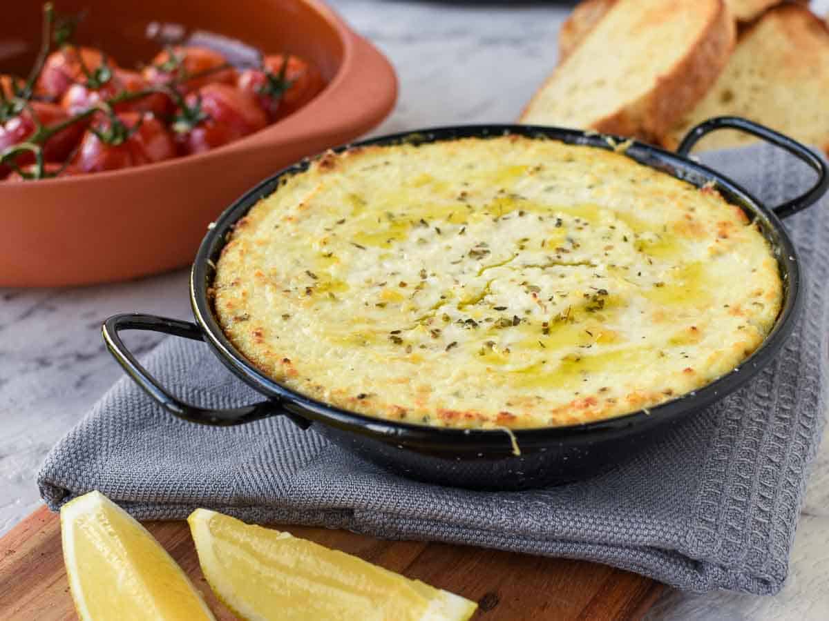 30 minutes. Serves 8. This Creamy Baked Ricotta Dip is full of lemon and garlic flavours. Serve with Roasted Tomatoes and crusty bread for a delicious, savory appetizer. creamy baked cheese in black enamel pan on grey cloth - roasted tomatoes in the background.