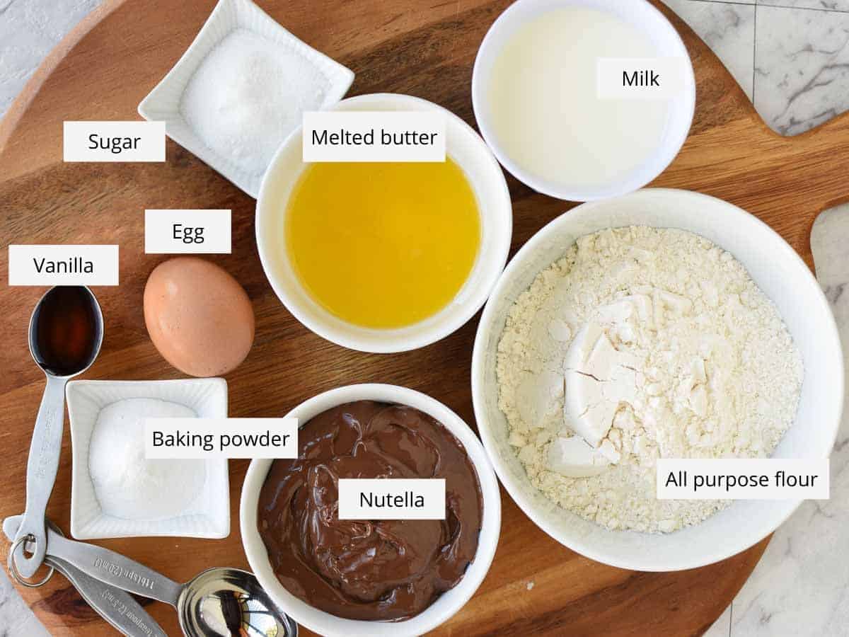 Makes 8 Nutella Stuffed Pancakes. Not just for breakfast, these Pancakes Stuffed with Nutella make a decadent snack or dessert. You'll love how easy this fluffy pancake recipe is! #pancakes #homemade #nutellapancakes #nutella Sugar, melted butter, milk , flour, nutella, baking powder, egg and vanilla in bowls viewed from above.