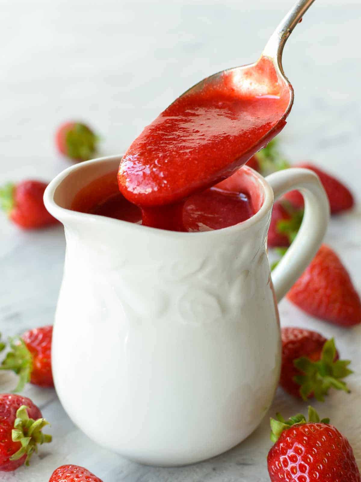 A homemade Strawberry sauce recipe using just 3 ingredients that is perfect for all desserts. You'll love using this strawberry topping for ice cream, pancakes or cheesecakes. #strawberries #dessert #sauce #strawberrysauce #strawberry #homemade a spoon full of strawberry sauce being poured into a white jug.