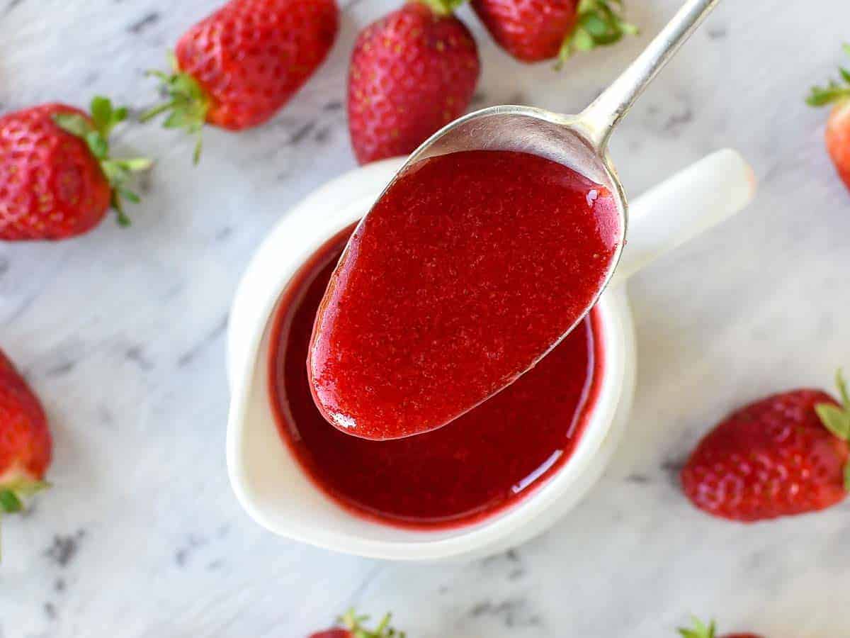 A homemade Strawberry sauce recipe using just 3 ingredients that is perfect for all desserts. You'll love using this strawberry topping for ice cream, pancakes or cheesecakes. #strawberries #dessert #sauce #strawberrysauce #strawberry #homemade a spoon full of strawberry sauce being poured into a white jug viewed from above.