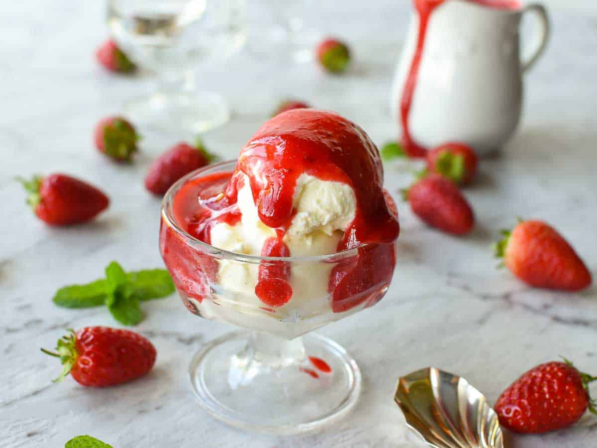 A homemade Strawberry sauce recipe using just 3 ingredients that is perfect for all desserts. You'll love using this strawberry topping for ice cream, pancakes or cheesecakes. #strawberries #dessert #sauce #strawberrysauce #strawberry #homemade glass bowl of vanilla ice cream with strawberry coulis on top; white jug in the background with a dribble of coulis on the jug.