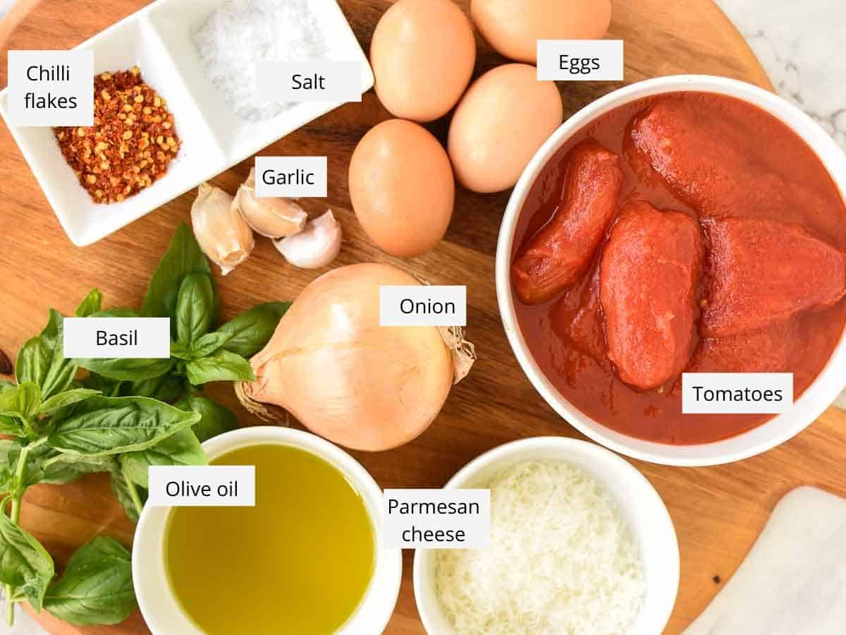 Eggs in Purgatory is an easy Italian dish you can prepare in less than 30 minutes. Eggs are poached in a spicy tomato sauce until the whites are set and the yolks are still creamy. Pure comfort food! #Italian #recipes #comfort #food #eggs #easyrecipe #eggsinpurgatory chilli flakes; salt; eggs; tinned tomatoes; grated parmesan cheese; oil; fresh basil; garlic on wooden board viewed from above.