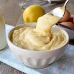 white bowl of pastry cream with a spoonful being poured in on pale blue cloth with bottle of milk, a lemon, eggs and a vanilla bean in the background