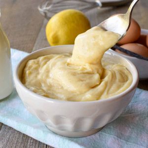 white bowl of pastry cream with a spoonful being poured in on pale blue cloth with bottle of milk, a lemon, eggs and a vanilla bean in the background
