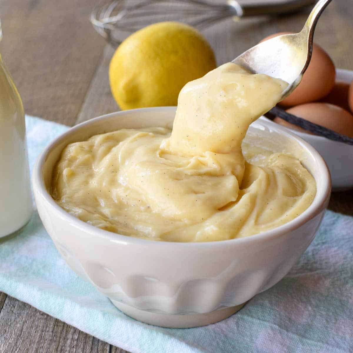 white bowl of pastry cream with a spoonful being poured in on pale blue cloth with bottle of milk, a lemon, eggs and a vanilla bean in the background