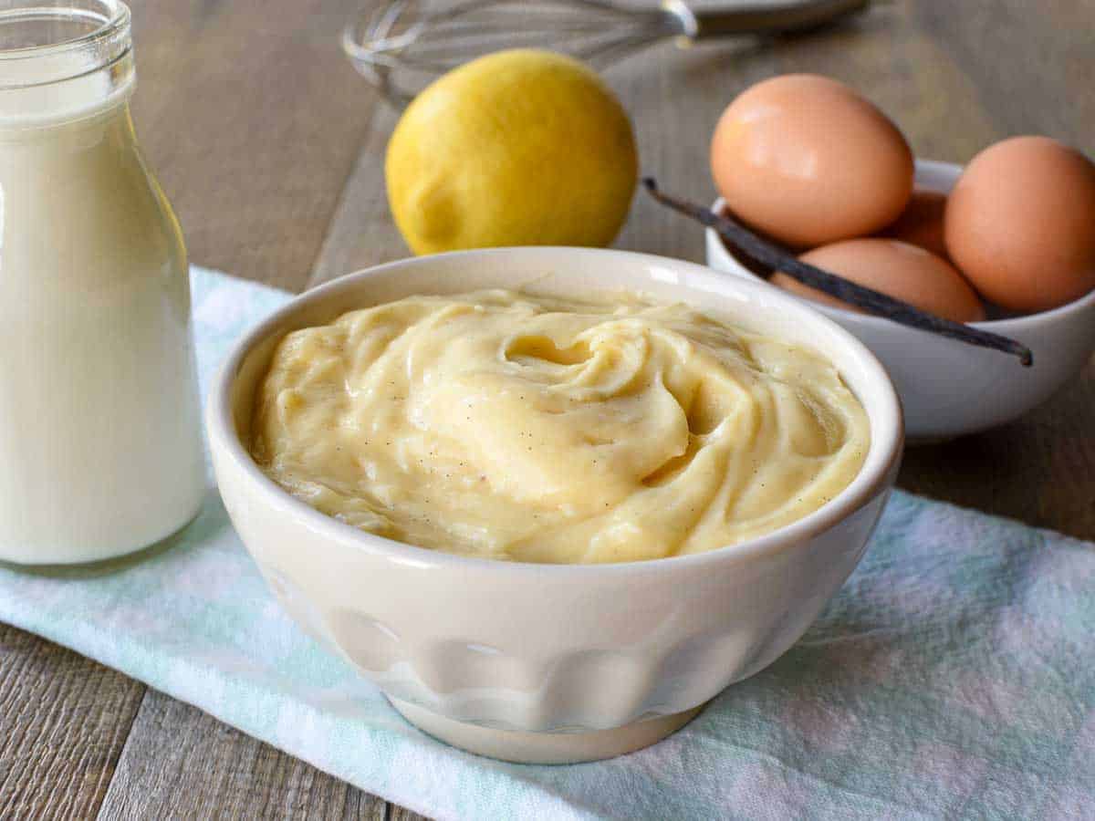 A thick and creamy Italian pastry cream that is perfect for pastries and desserts. This Italian version of creme patissiere has a delicate lemon and vanilla flavour and can be adapted to make other flavours such as chocolate or coffee. #italianpastry cream #crema pasticcera #cremepat #cremepatissiere #pastrycream #custard #vanillapastrycream white bowl of pastry cream on pale blue cloth with bottle of milk, a lemon, eggs and a vanilla bean in the background.