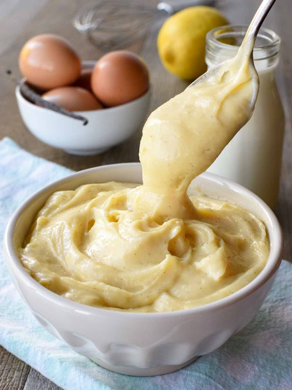 A thick and creamy Italian pastry cream that is perfect for pastries and desserts. This Italian version of creme patissiere has a delicate lemon and vanilla flavour and can be adapted to make other flavours such as chocolate or coffee. #italianpastry cream #crema pasticcera #cremepat #cremepatissiere #pastrycream #custard #vanillapastrycream white bowl of pastry cream with a spoonful being poured in on pale blue cloth with bottle of milk, a lemon, eggs and a vanilla bean in the background.