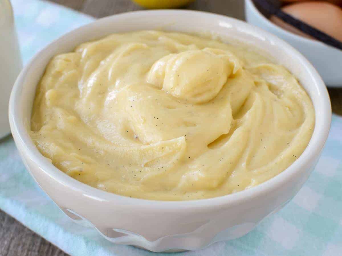 A thick and luscious cream that is perfect for pastries and desserts. This Italian version of creme patissiere has a delicate lemon and vanilla flavour and can be adapted to make other flavours such as chocolate or coffee. #italianpastry cream #crema pasticcera #cremepat #cremepatissiere #pastrycream #custard #vanillapastrycream white bowl of pastry cream on pale blue cloth.