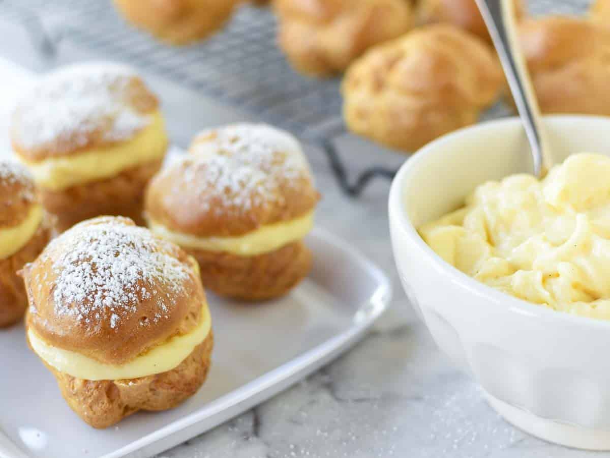 How to make Cream puffs with this step by step recipe. This classic Italian pastry that is not difficult to make and you'll love the pastry cream filling. #creampuffs #creampuffrecipe #dessert #pastry #pastries #italianrecipe pastries filled with custard on white plate, white bowl of custard and unfilled pastries in the background