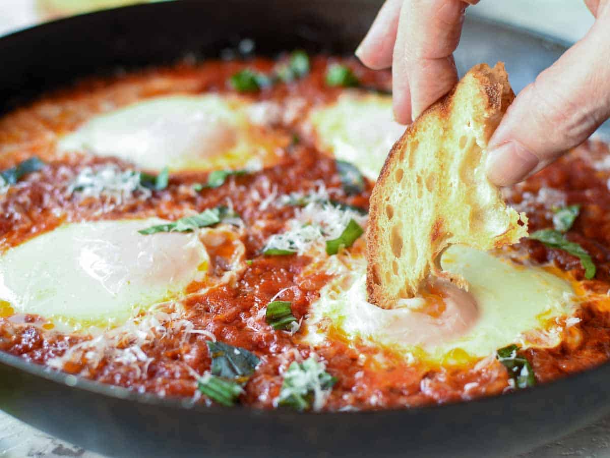 An easy Italian egg dish you can prepare in less than 30 minutes. Eggs are poached in a spicy tomato sauce until the whites are set and the yolks are still creamy. Pure comfort food! #Italian #recipes #comfort #food #eggs #easyrecipe #eggsinpurgatory Poached eggs in tomato sauce in black pan, fingers holding toasted bread dipping into egg.