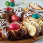 close up of baked sweet bread decorated with red, blue, yellow and green eggs and drizzle of icing and a few slice cut to show inside of bread. At the rear is two slice of the bread on a white plate. Viewed from front