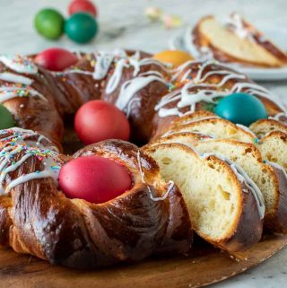 close up of baked sweet bread decorated with red, blue, yellow and green eggs and drizzle of icing and a few slice cut to show inside of bread. At the rear is two slice of the bread on a white plate. Viewed from front