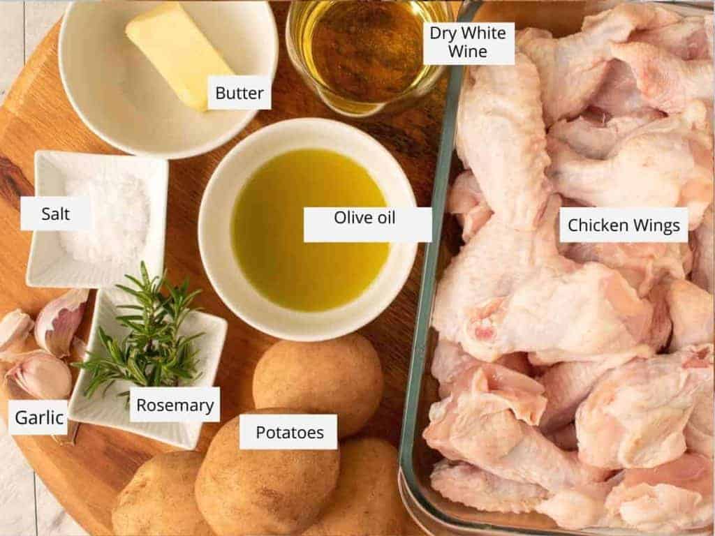 Butter, dry white wine, raw chicken wings, potatoes, rosemary, garlic, salt and olive oil in bowls and dishes on a wooden board