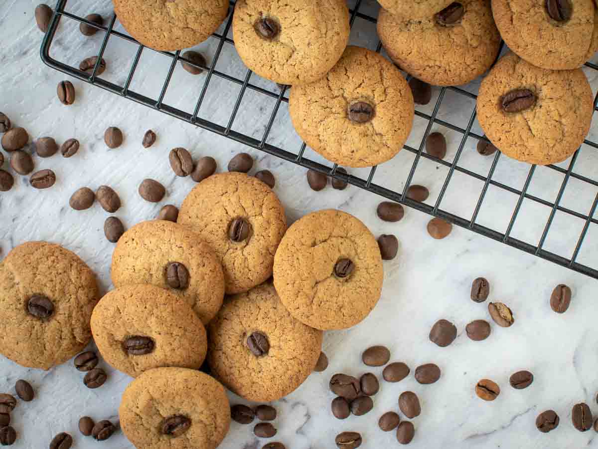 Cookies on black wire rack and more on marble surface with coffee beans scattered around viewed from above.