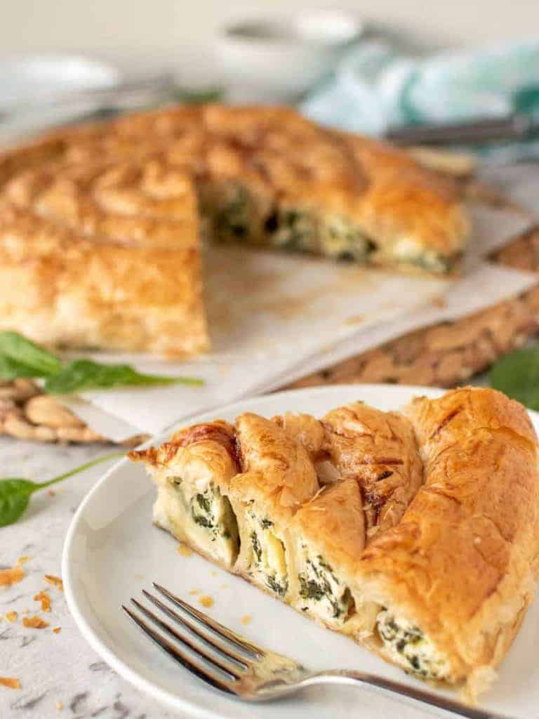 slice of spinach ricotta filo pie on white plate with remaining pie in the background