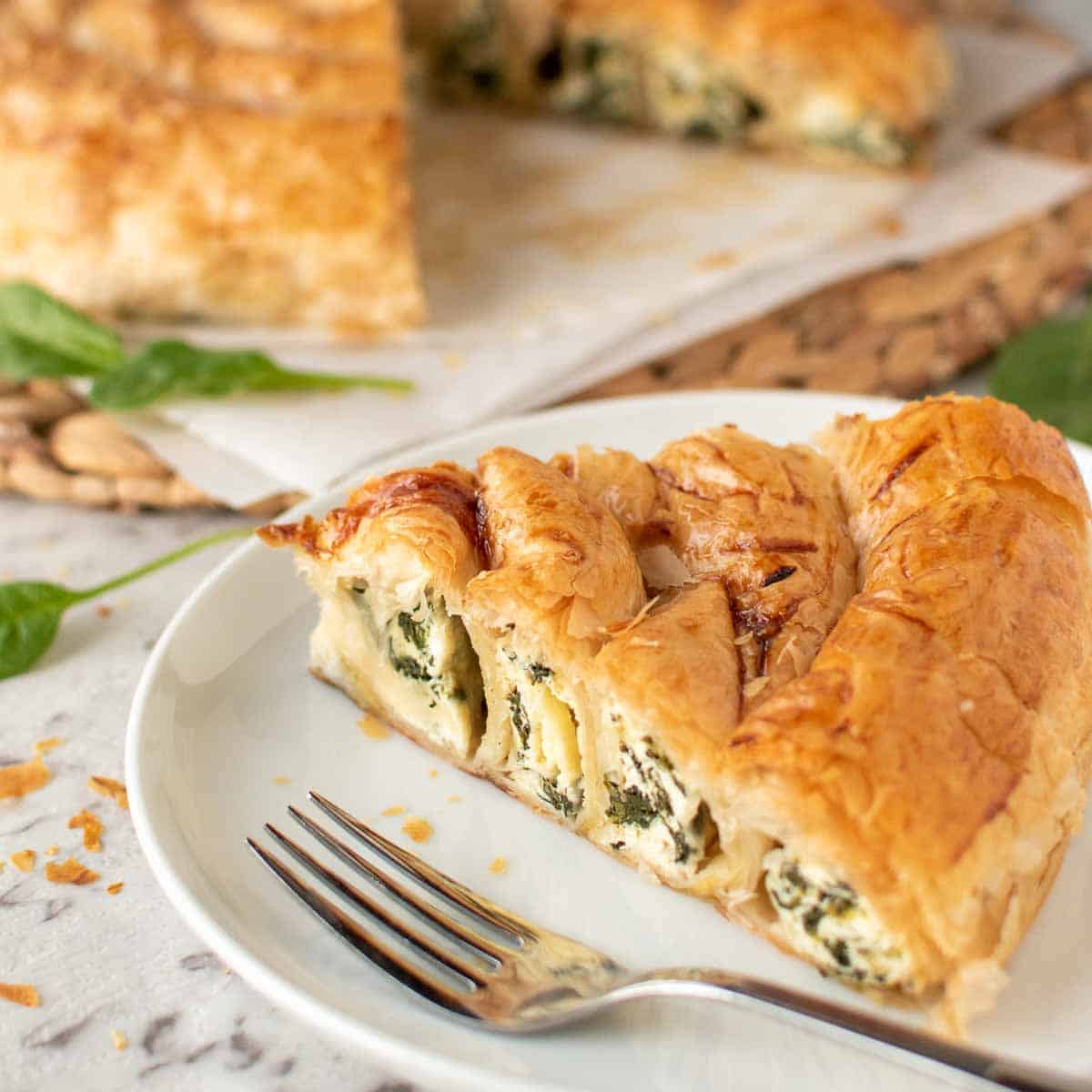slice of spinach ricotta filo pie on white plate with remaining pie in the background