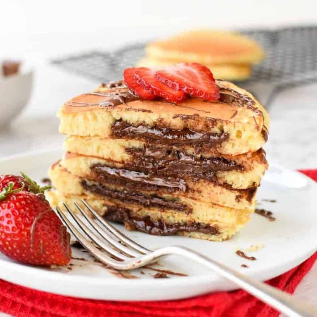 five stacked nutella filled pancakes on white plate sliced in half showing the nutella oozing out