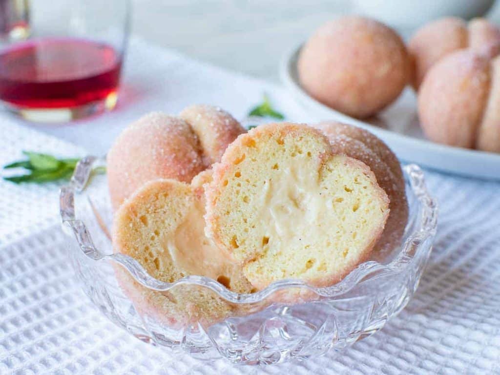 A cookie resembling a peach cut in half revealing pastry cream center in a glass bowl with more whole cookies.