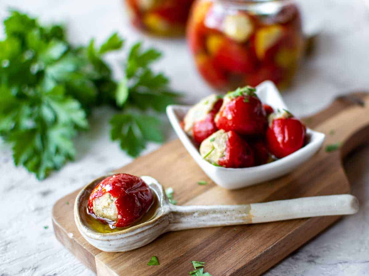 one stuffed cherry pepper in a spoon and pile of stuffed cherry peppers in a white dish with bottles filled in the background