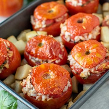 eight baked tomatoes stuffed with rice in black baking pan with cubed potatoes.