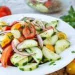 close up of cucumber tomato onion salad on white plate with tomatoes and parsley in the background