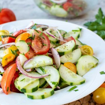close up of cucumber tomato onion salad on white plate with tomatoes and parsley in the background