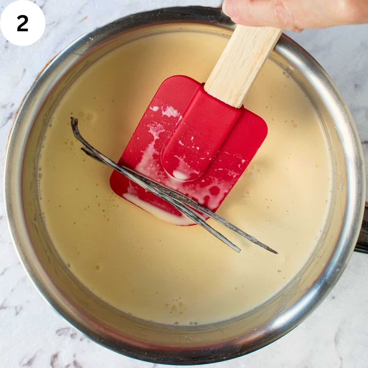 A split vanilla bean being lifted with a silicone spatula in a saucepan of cream.