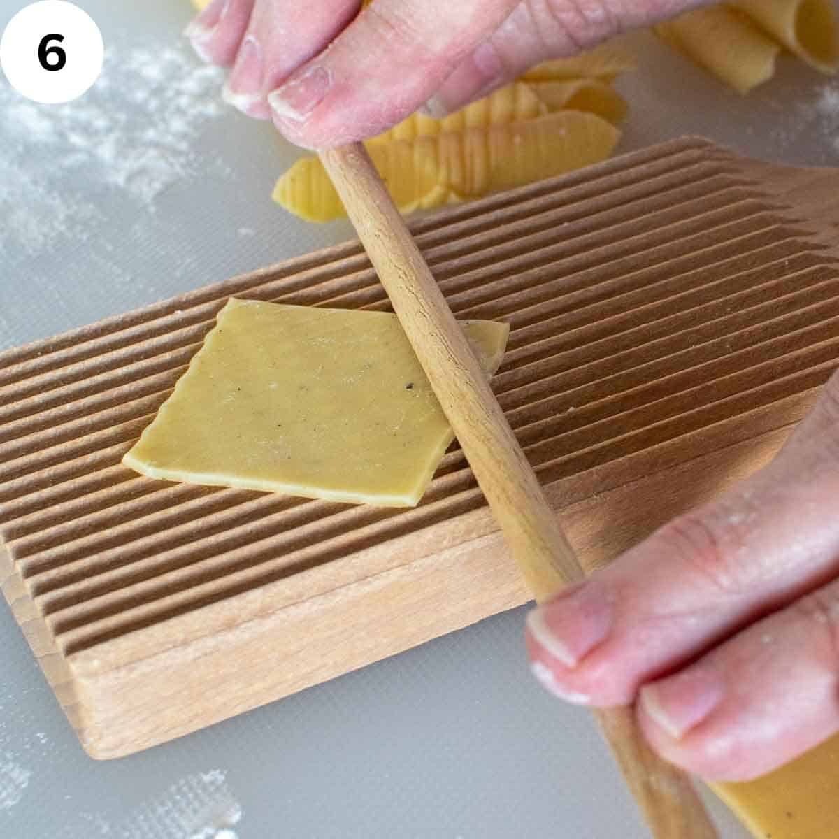 A small square of pasta dough on a ridged board with a wooden dowel across one end of the square.