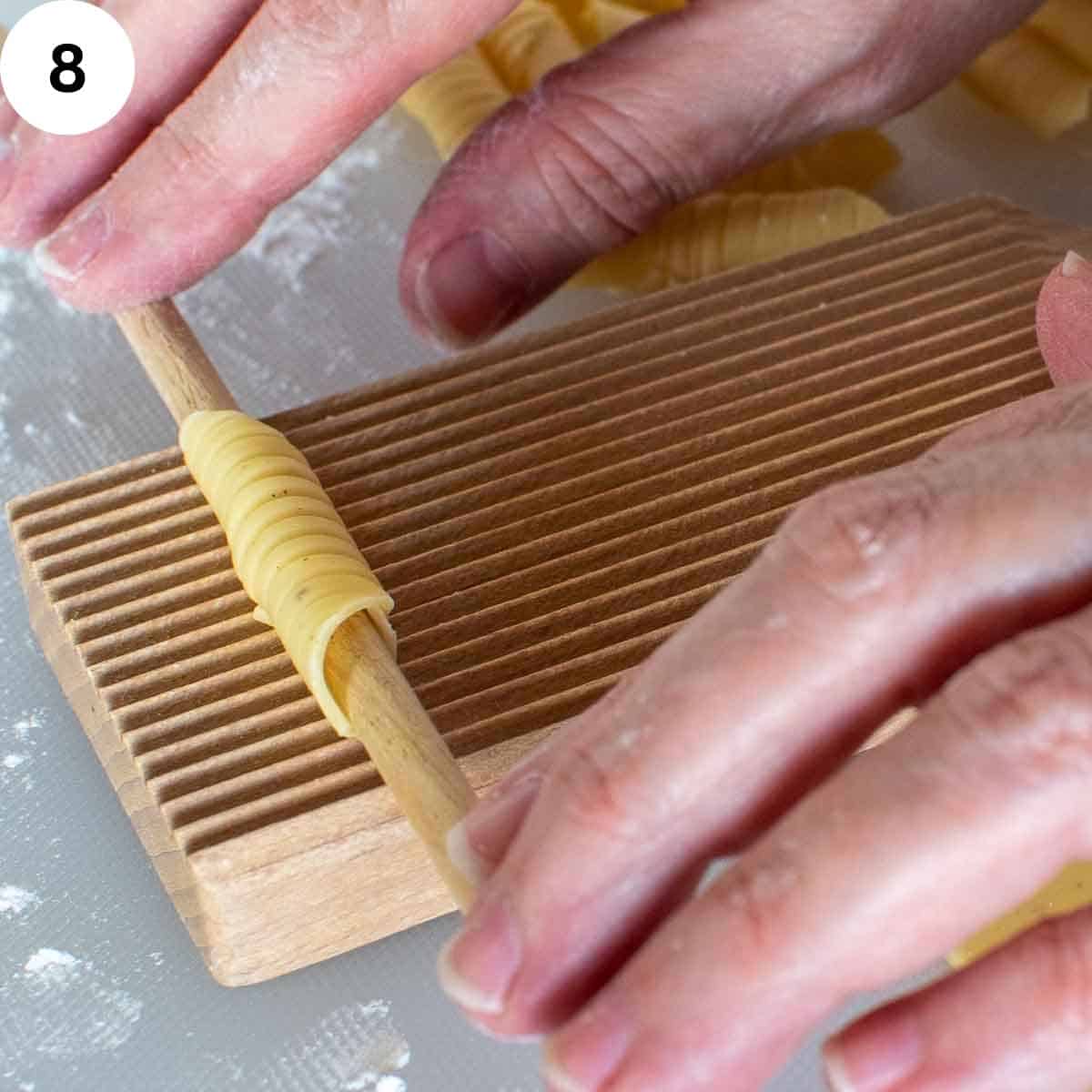 Tubular shaped pasta dough with a wooden dowel through the middle on a ridged board.
