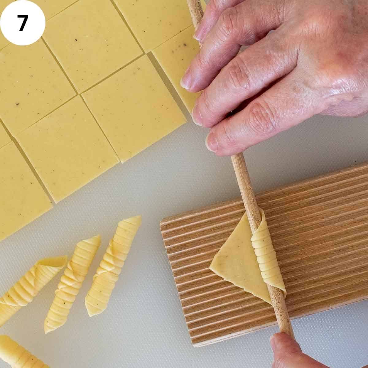 A small square of pasta on a ridged board being rolled around a wooden dowel.