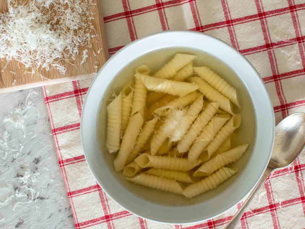 Bowl of broth with tubular shaped pasta in the broth checked cloth and grated parmesan cheese on wooden board viewed from above.