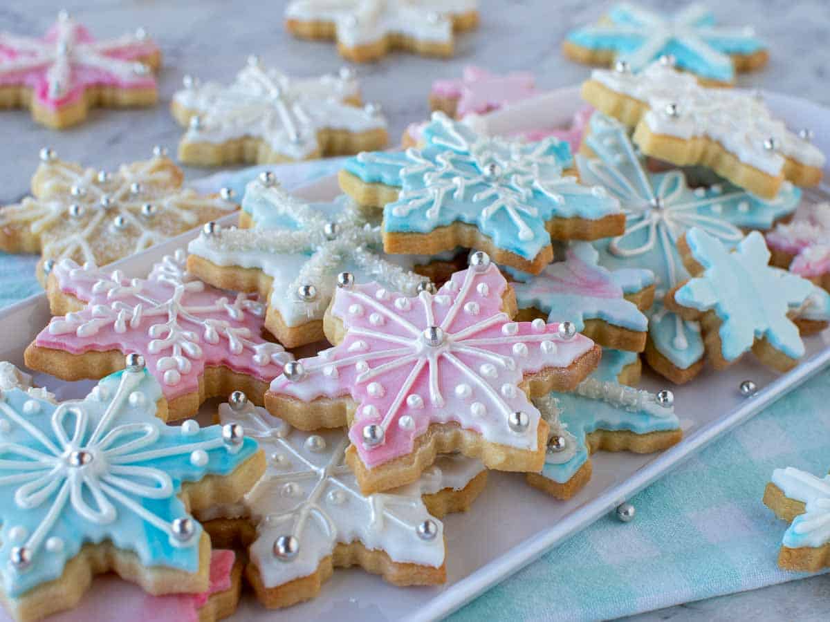 Pink, white and blue decorated sugar cookies in the shape of snowflakes on white oblong plate.