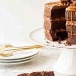 slice of three layer chocolate cake with chocolate frosting and caramel with whole cake in the background on a white stand.