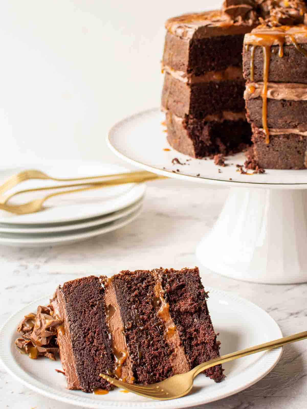 A slice of Chocolate Cake with Caramel on a plate; whole cake in the background.
