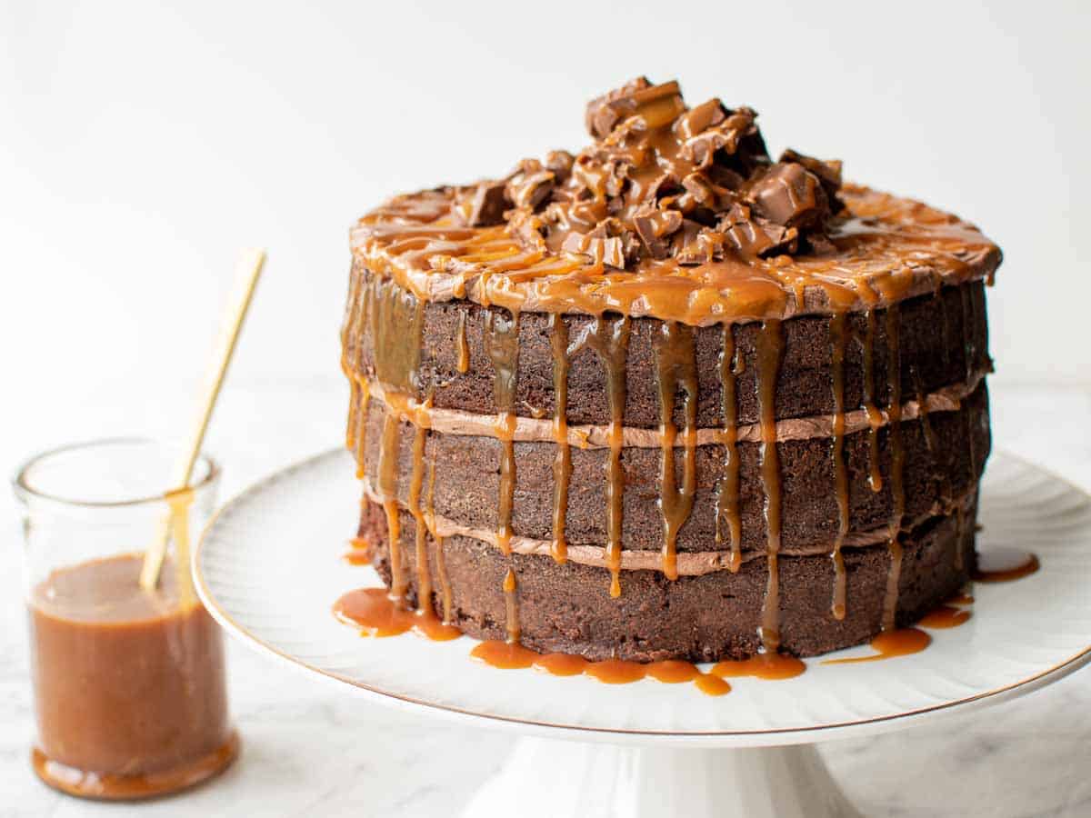three layer chocolate cake with chocolate frosting and dripping caramel on a white cake stand with jar of caramel in the background.