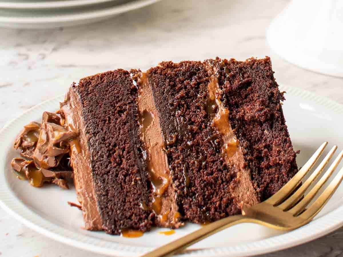 Close up of a slice of three layered chocolate cake with frosting on a plate with a fork.