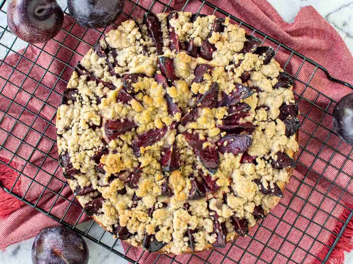 Cake topped with slices plums and streusel on black wire rack viewed from above.