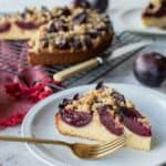 Wedge of plum cake on white plate with gold fork and whole cake in the background.