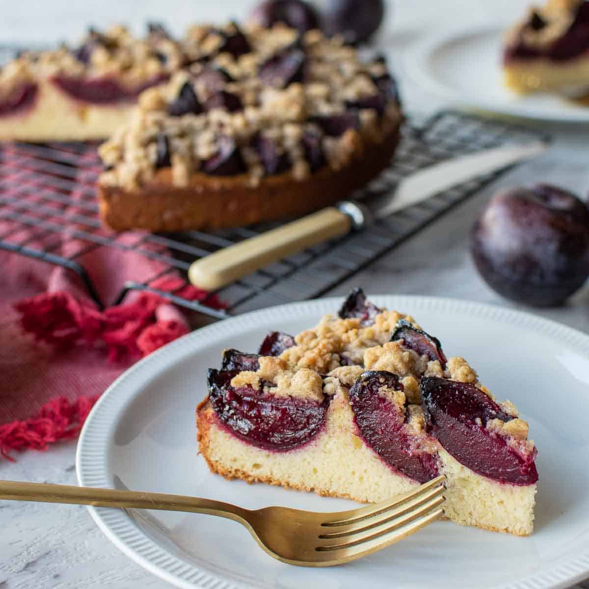 Wedge of plum cake on white plate with gold fork and whole cake in the background.