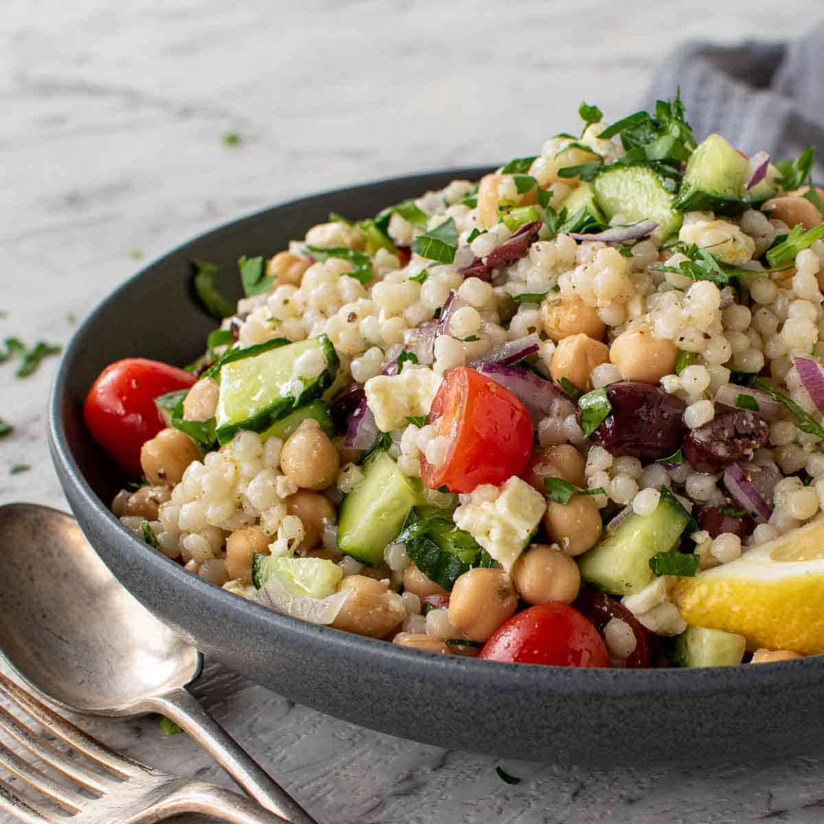 bowl of couscous salad with fork and spoon in the front and grey towel in the background.