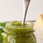 Image with text. Text reads "How to make nut free pesto easy and homemade. Image is glass jar of basil pesto with a spoon pushed into the pesto, fresh basil in the background.