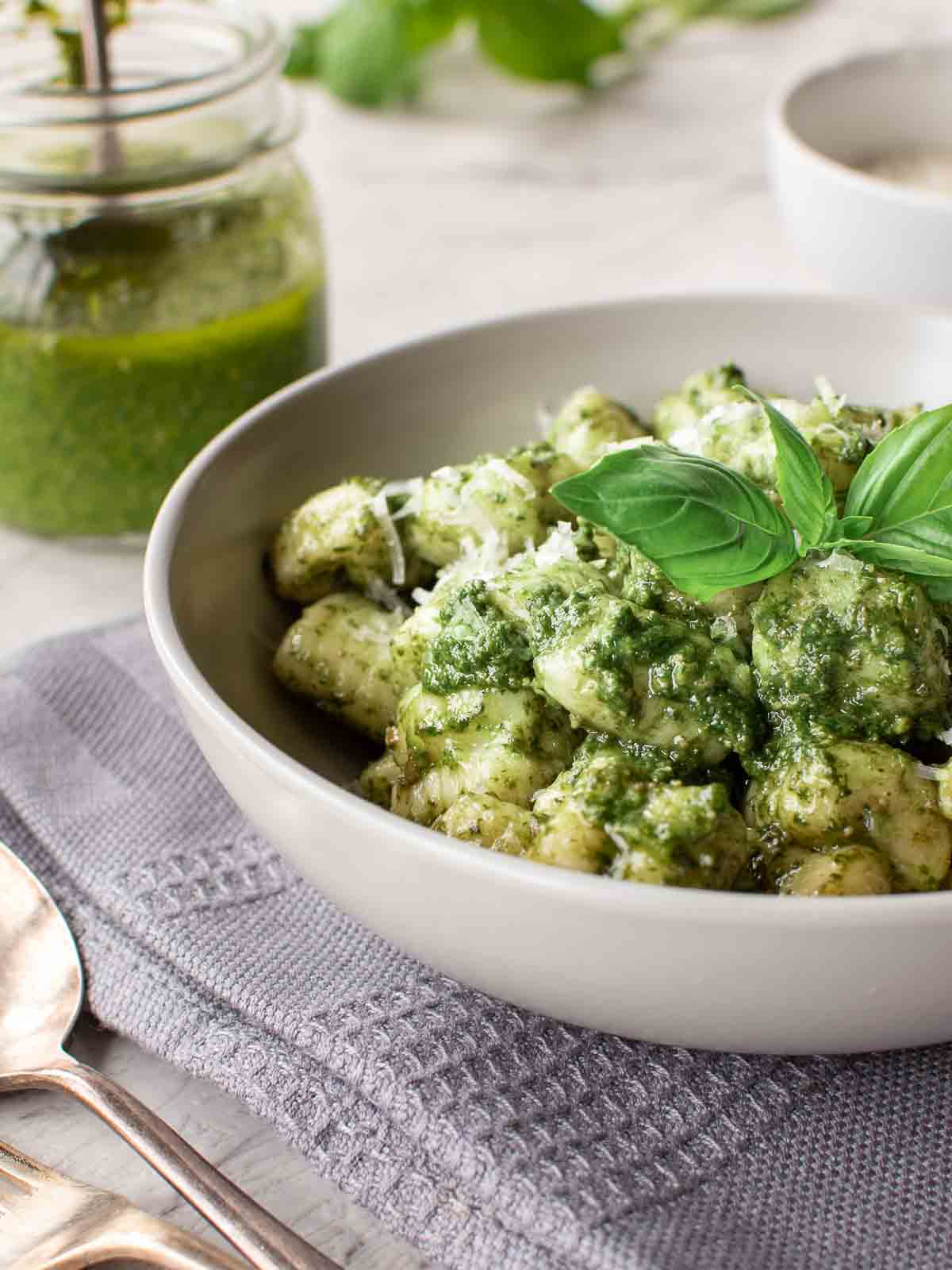 A bowl this pesto gnocchi recipe on a grey cloth with jar of pesto in the background.