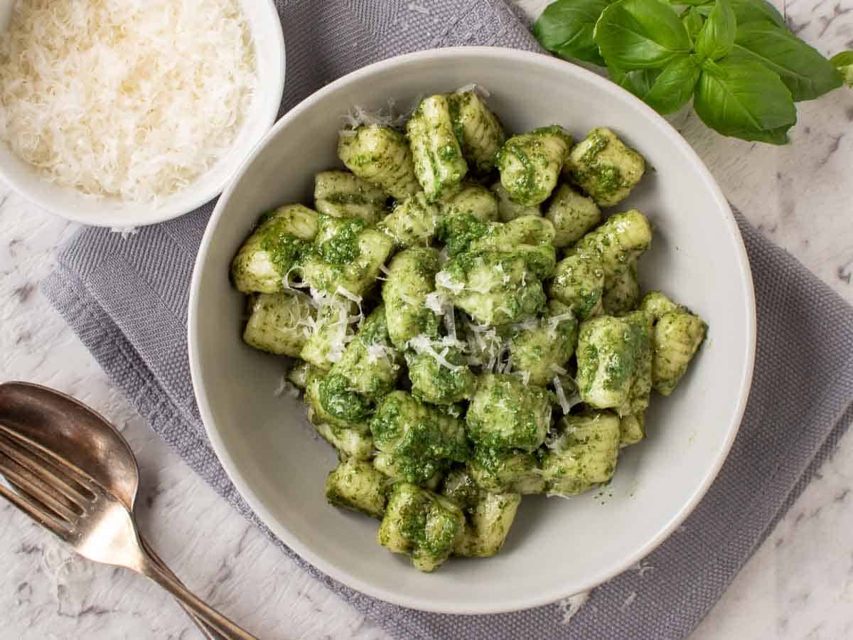 A bowl of gnocchi on a grey cloth with bowl of grated parmesan on the side viewed from above.