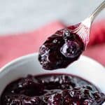 image with text. text reads 'quick and easy blueberry compote". image is blueberry compote in a white bowl with a spoon full lifted out.