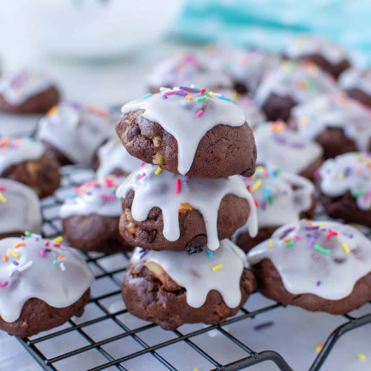 close up of three chocolate cookies with white frosting and sprinkles stacked with extra cookies around all on a wire rack.