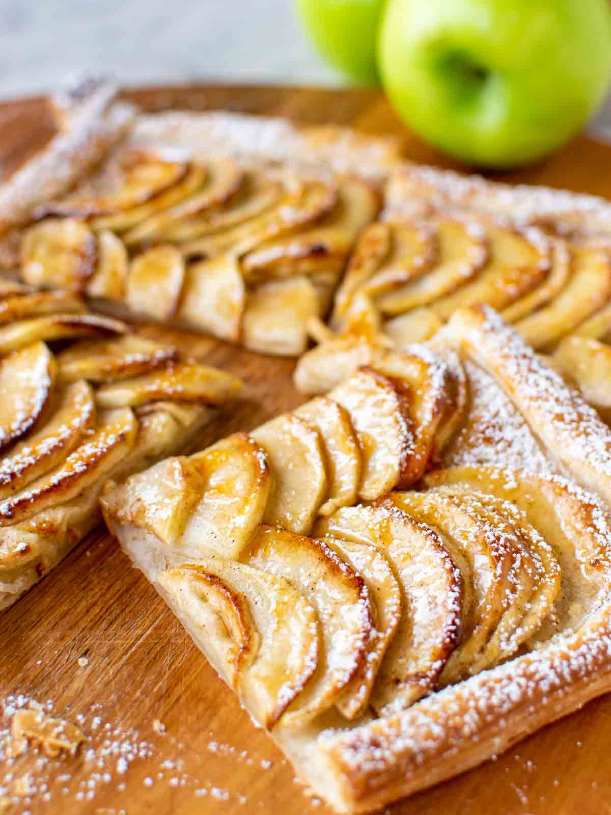 Slices of puff pastry apple tart on wooden board, green apple in the background.