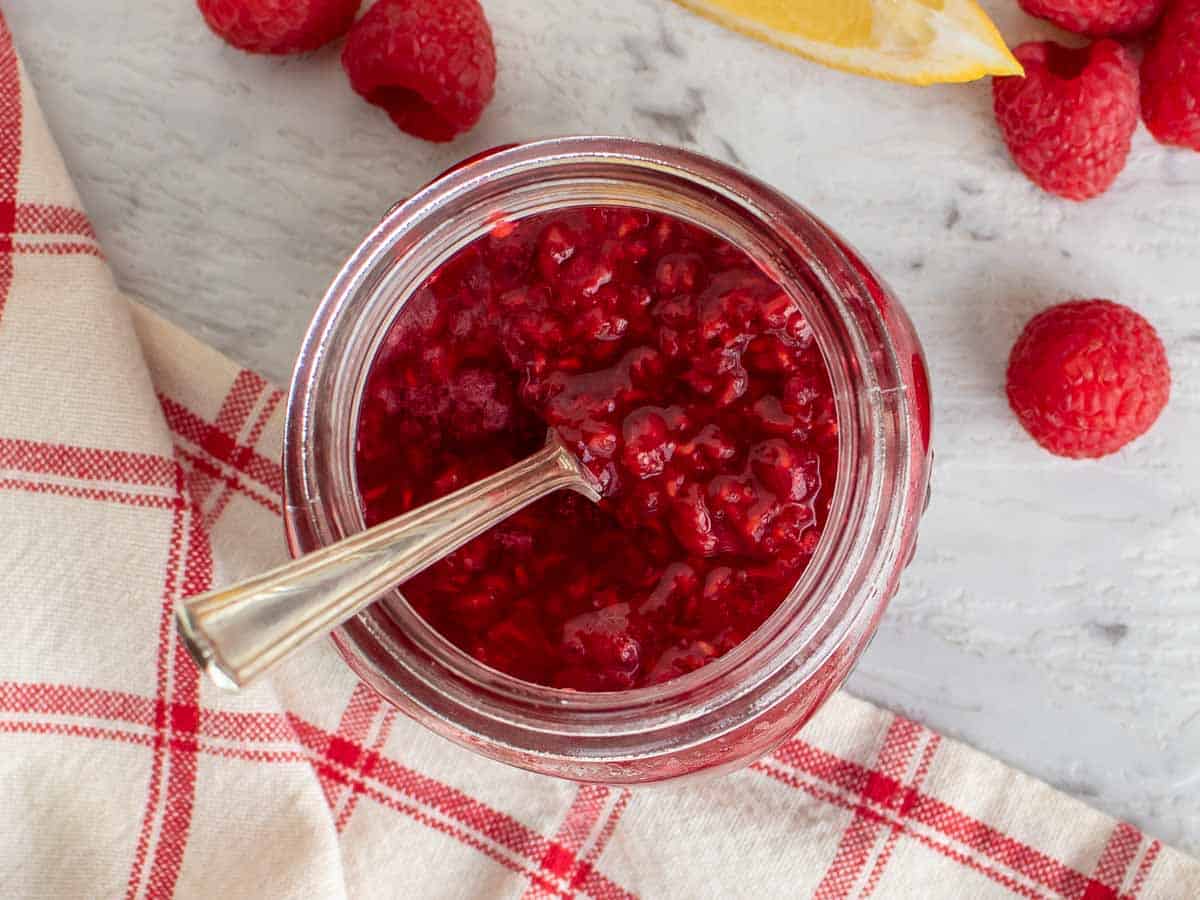Raspberry sauce in a glass jar with spoon insert viewed from above.